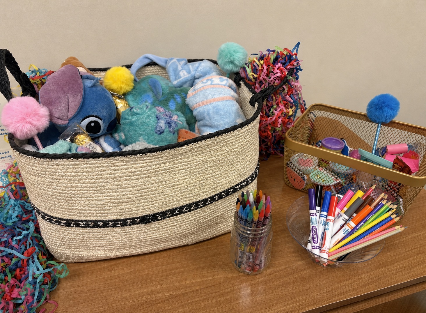 Woven basket filled with stuffed animals and colorful tactile items beside jars of crayons, markers, and pencils